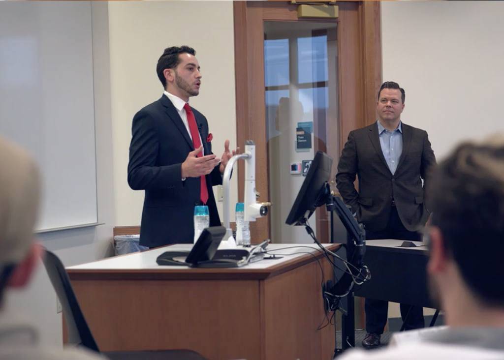 A male student stands and listens as a male instructor, dressed in a suit, speaks animatedly.